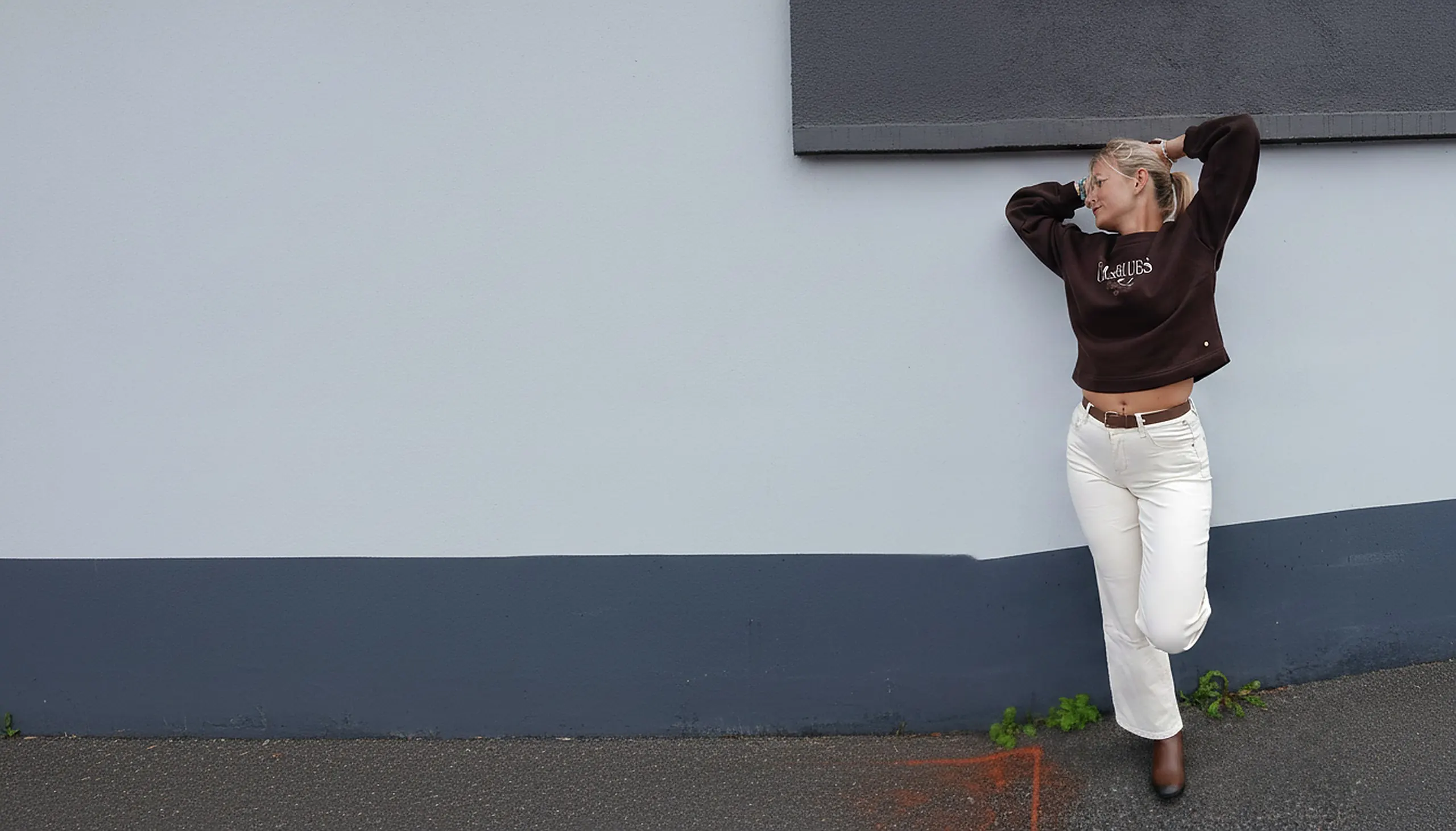 Laurie porte un sweat Red Button et un pantalon à ceinture marron devant un mur clair