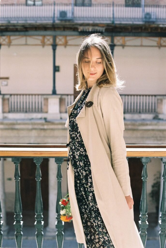 Femme en robe midi sombre à petites fleurs et trench beige ouvert, sur une galerie ensoleillée