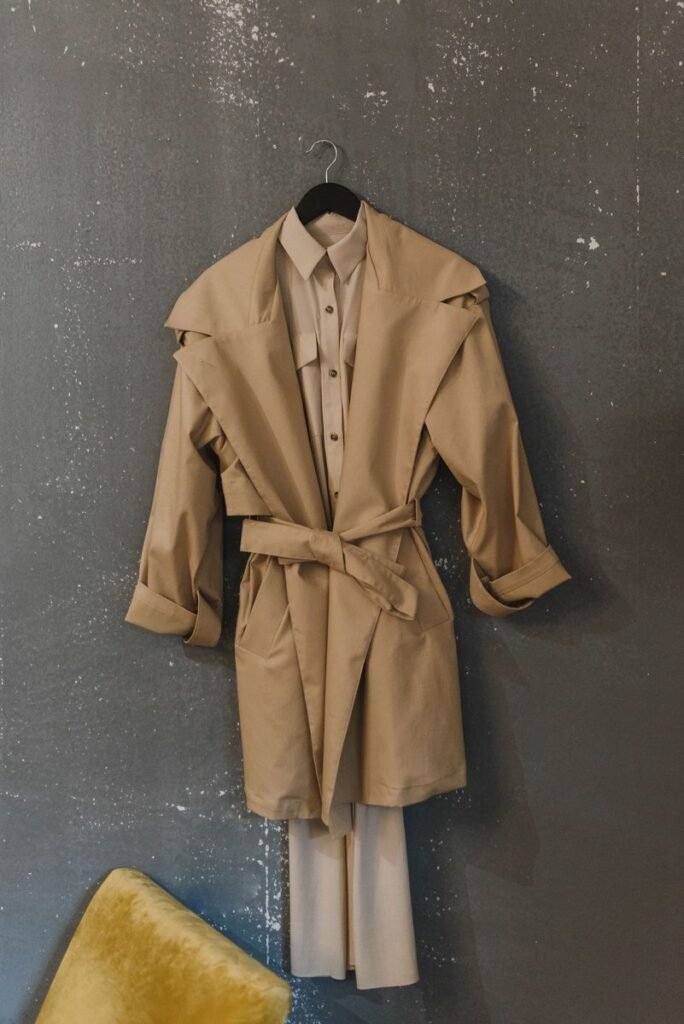 Trench beige ceinturé suspendu à un cintre sur une robe chemise, contre un mur gris