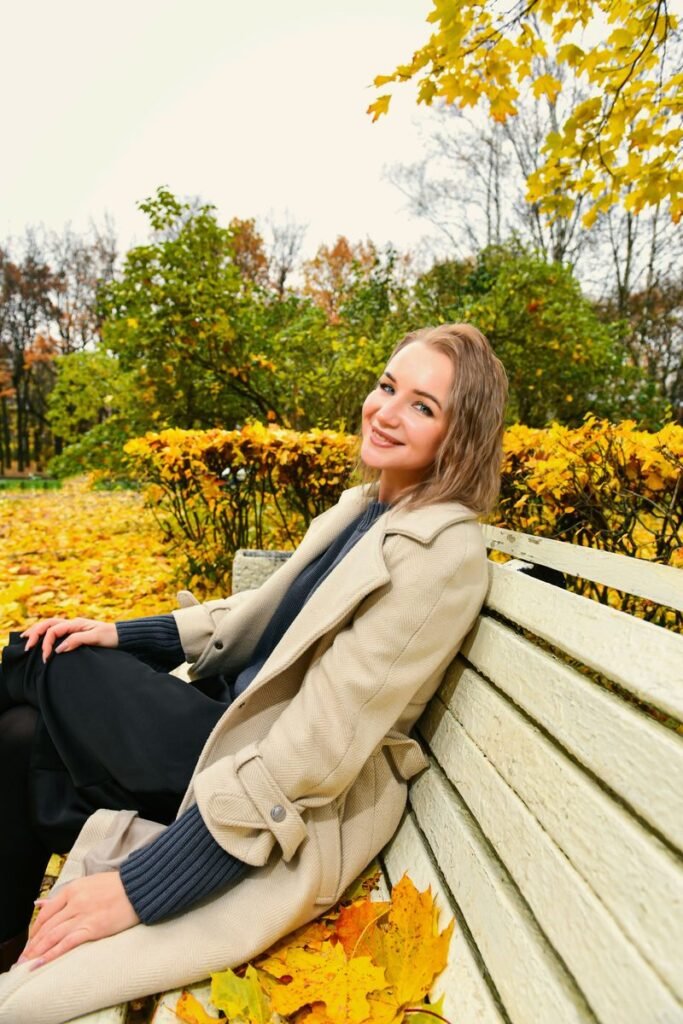 Femme souriante en trench beige sur un pull bleu marine, assise sur un banc parmi les feuilles d’automne