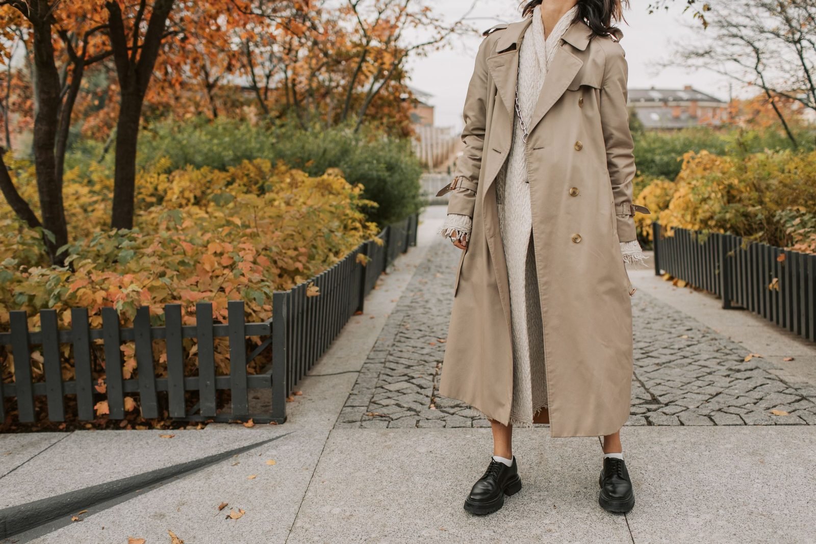 Trench beige long ouvert sur une robe en maille crème et derbies noires, dans une allée aux feuillages d’automne