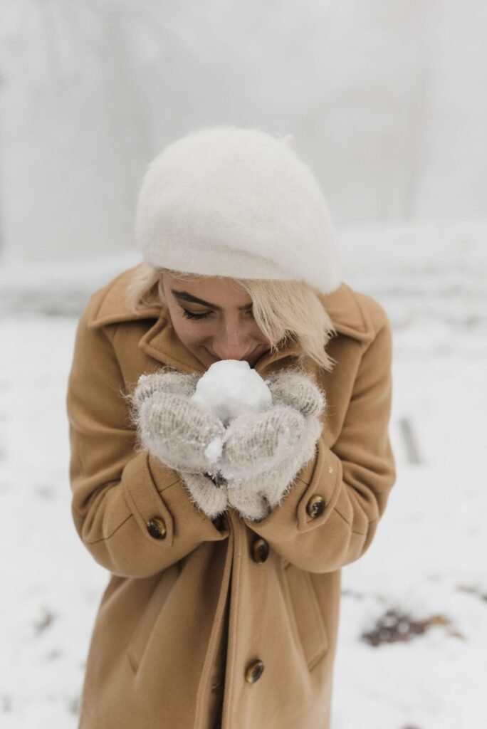 Femme en manteau en laine camel boutonné, col relevé, béret blanc et moufles en laine dans la neige