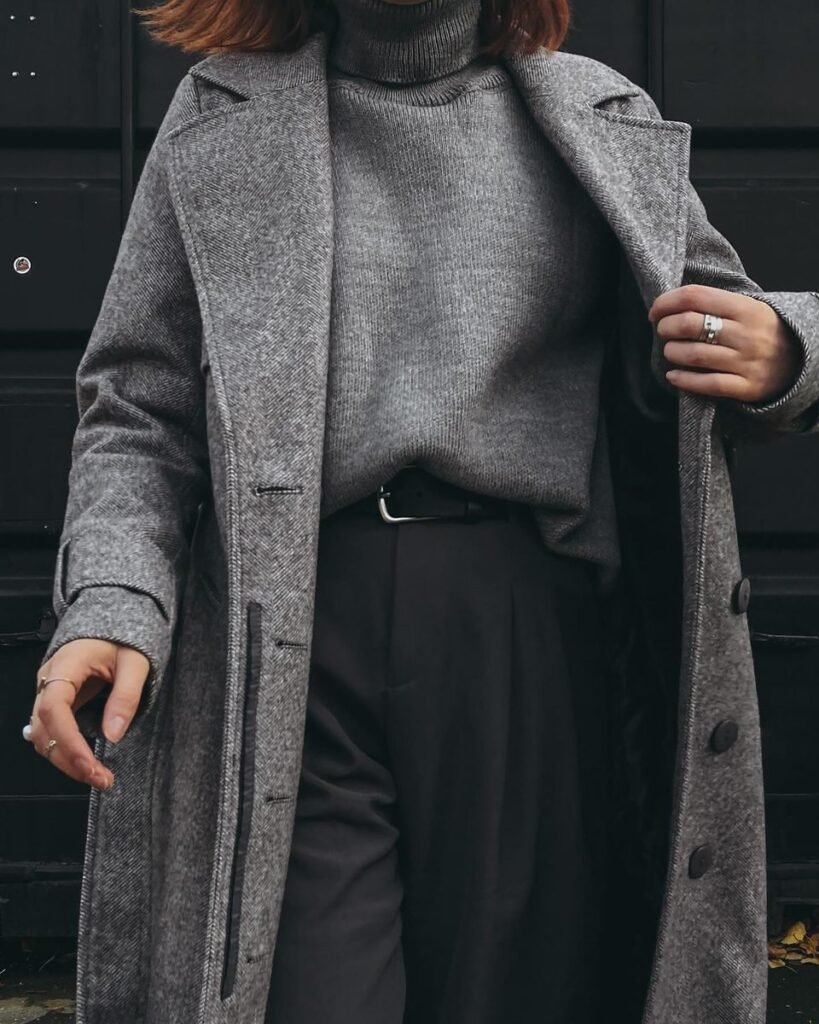 Pull col roulé gris rentré dans un pantalon noir à ceinture, manteau long gris chiné ouvert par-dessus